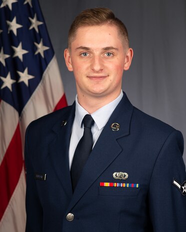Airman 1st Class poses in uniform for an official photo in front of the American flag.