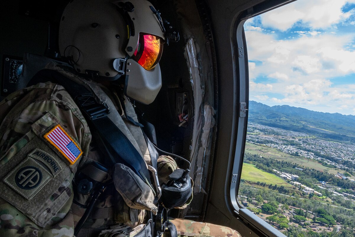 A uniformed soldier wearing goggles looks from a helicopter as it flies over land during daylight.