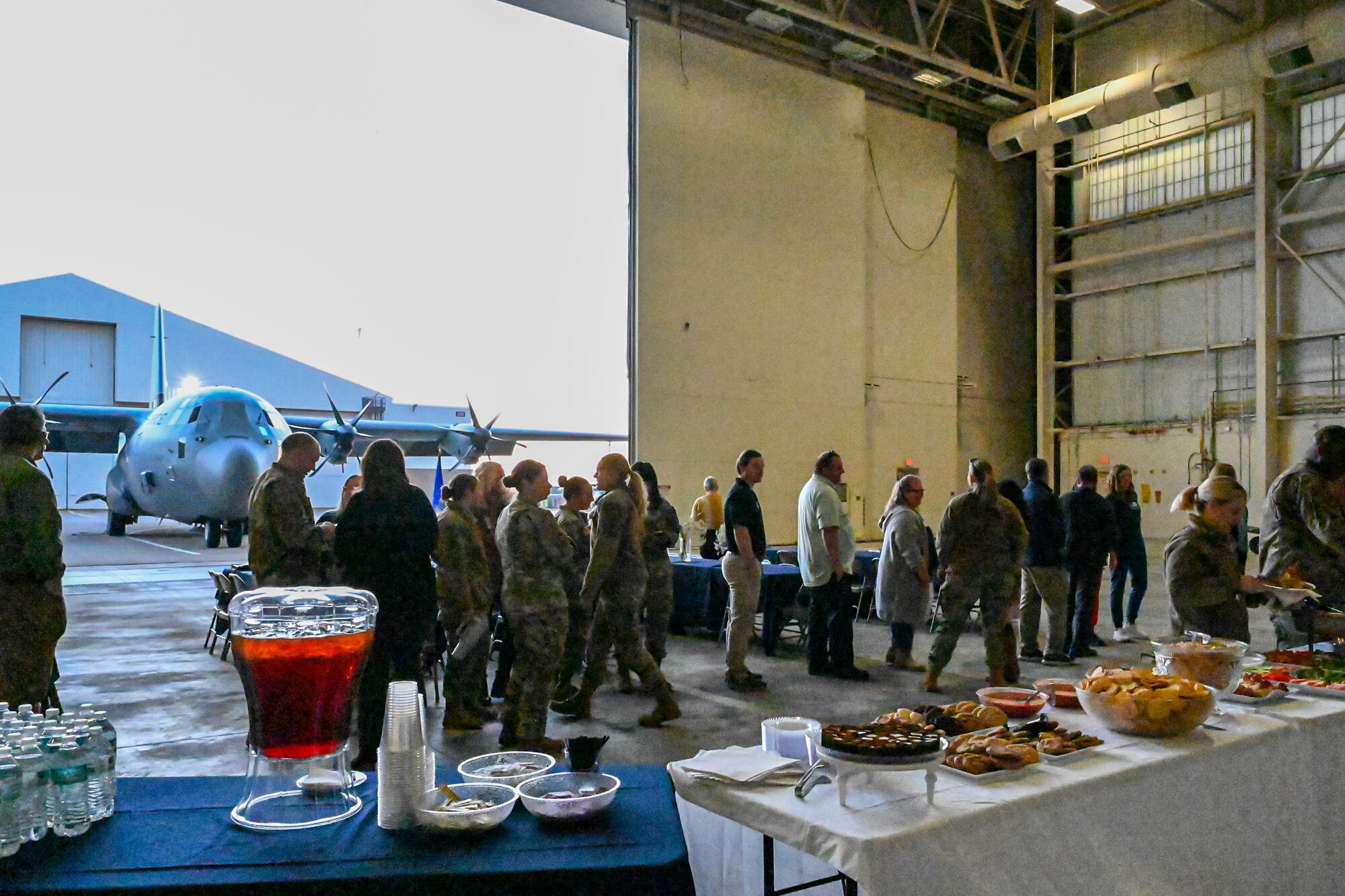 U.S. Airmen and civilians stand in a line to a buffet inside a hangar with a C-130J aircraft in the background.