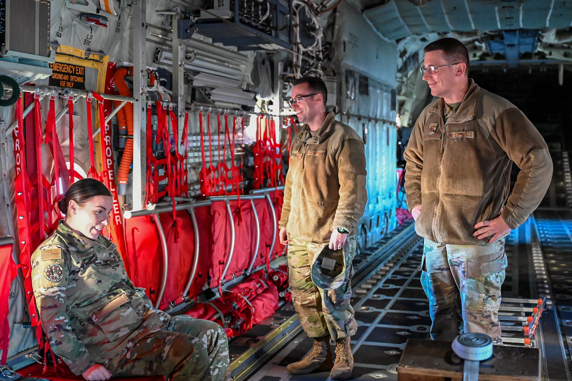 Three U.S. Airmen sit and stand inside a C-130J aircraft.