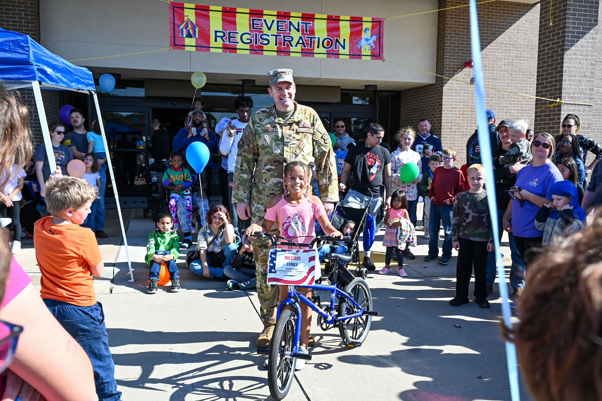 A U.S. Airman poses with a child and a bicycle for a photo within a crowd of people.