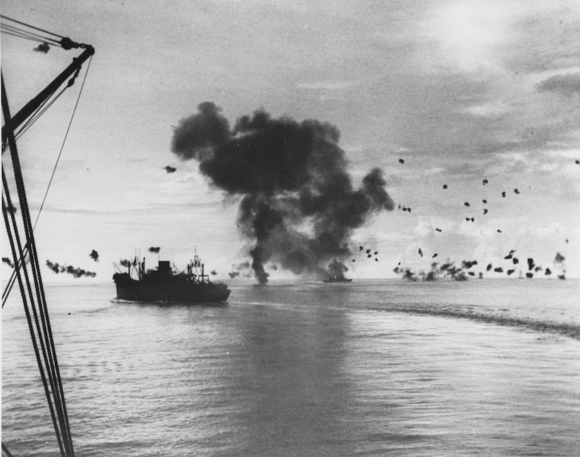 A large ship sits in water as another ship burns heavily in the distance.