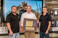 Three people pose for a photo in front of a large tablet with a military vehicle on it. The person at center holds a plaque listing the names of Tobyhanna Army Depot's Purple Heart recipients.