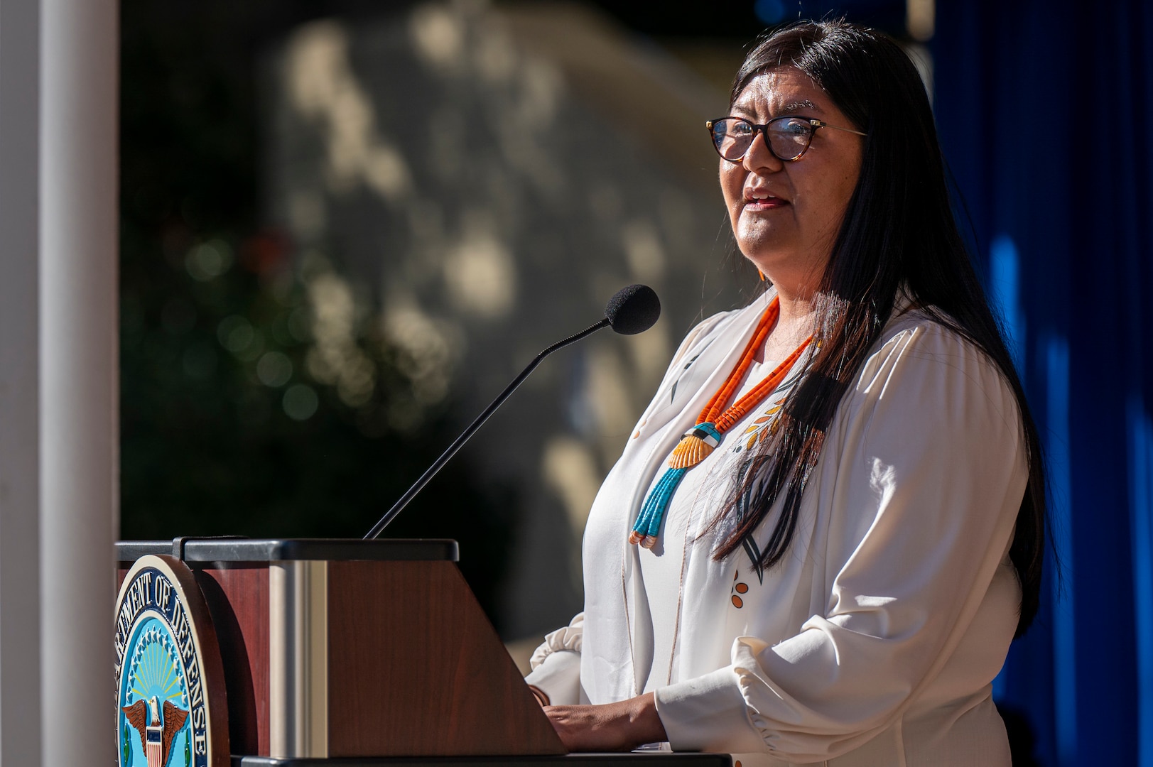 A civilian wearing a shell necklace speaks at a lectern outdoors.
