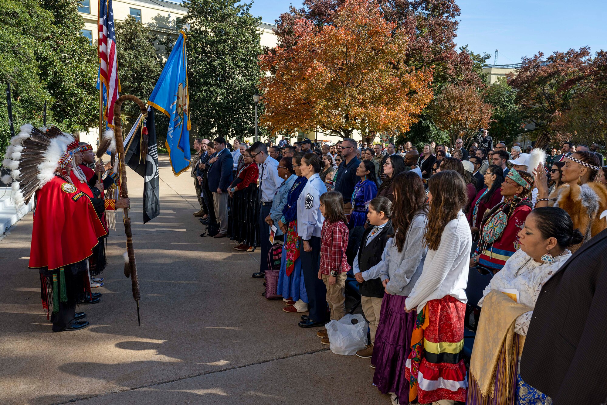People wearing traditional Native American clothing stand and hold flags while facing a standing audience outdoors.