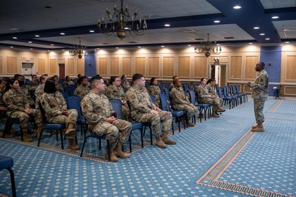 Group of Airmen sitting in chairs on left, Chief Williams standing on right.