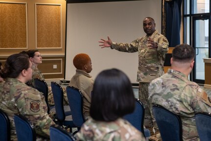 Chief Williams standing with arms open, addressing audience of Airmen sitting down.