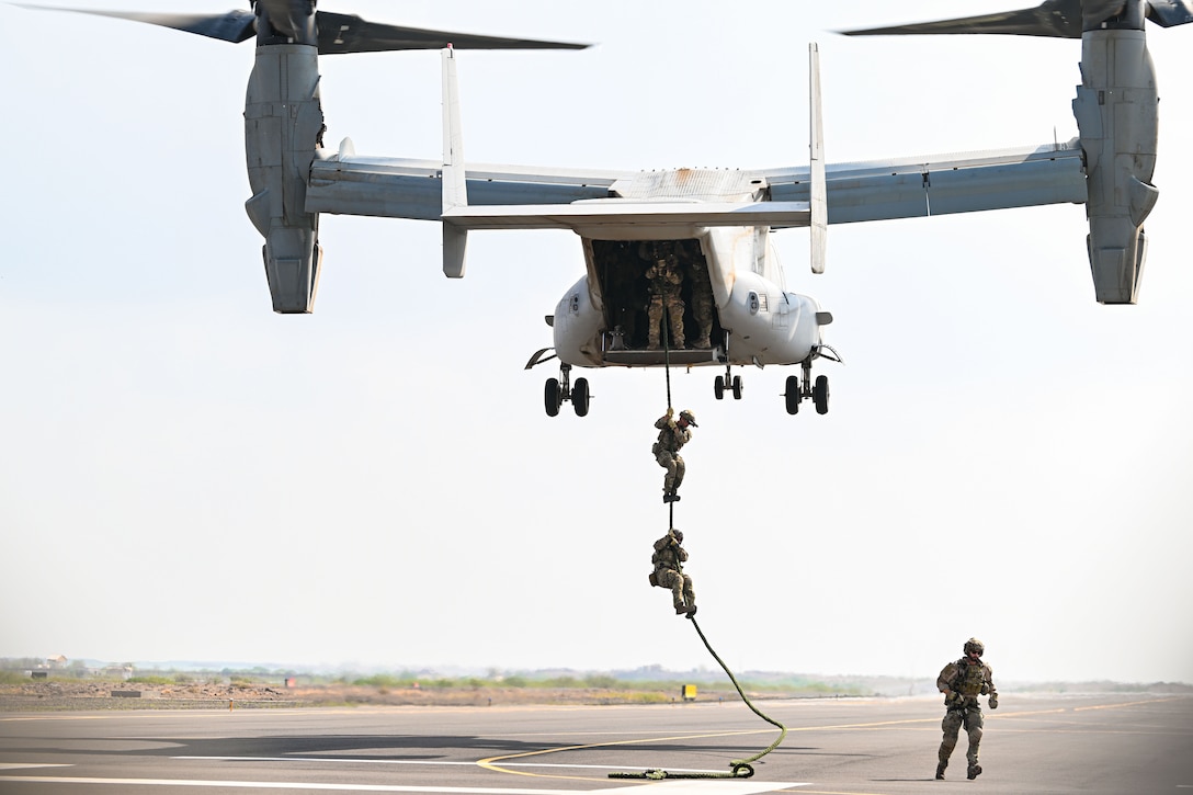 Airmen hold onto a rope dangling from a military aircraft hovering low over the ground.