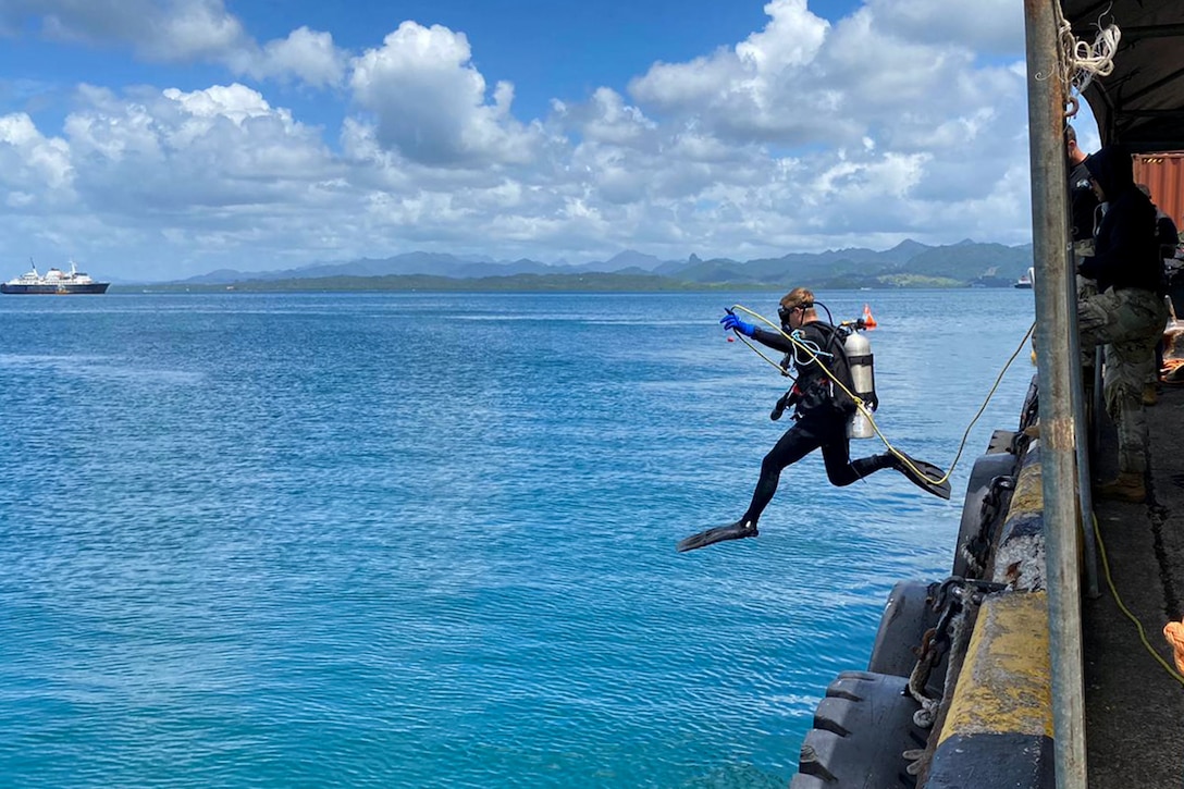 A person wearing a wet suit, goggles and an oxygen pack jumps from a pier into open water during daylight.