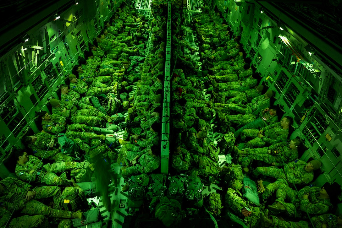 Dozens of soldiers sit in the cargo area of a military aircraft, illuminated by a green light.