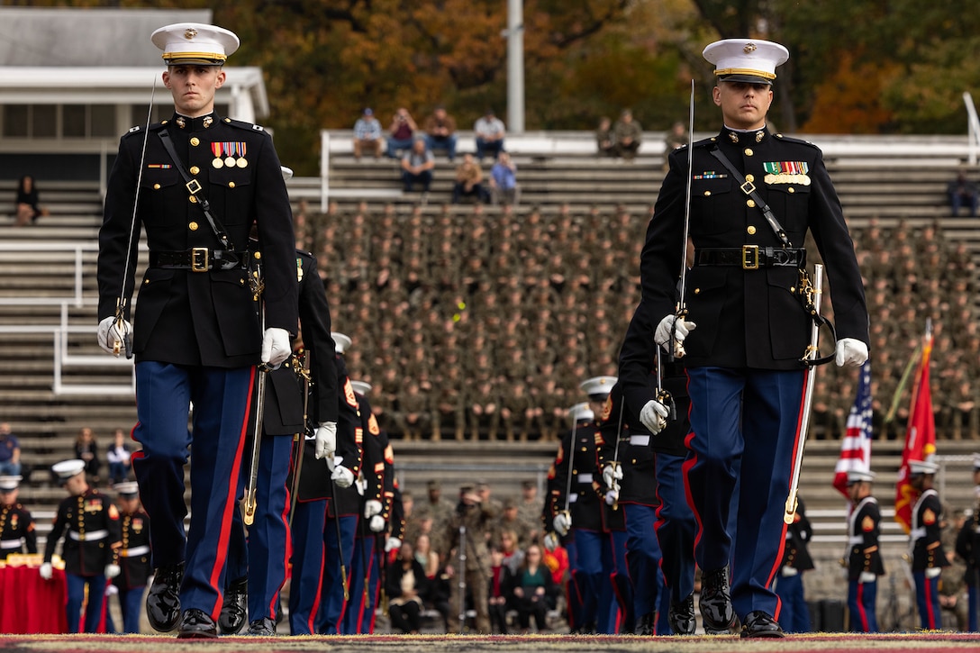 Marines in ceremonial dress, carrying swords, march in formation on a field as others sit in bleachers behind them.