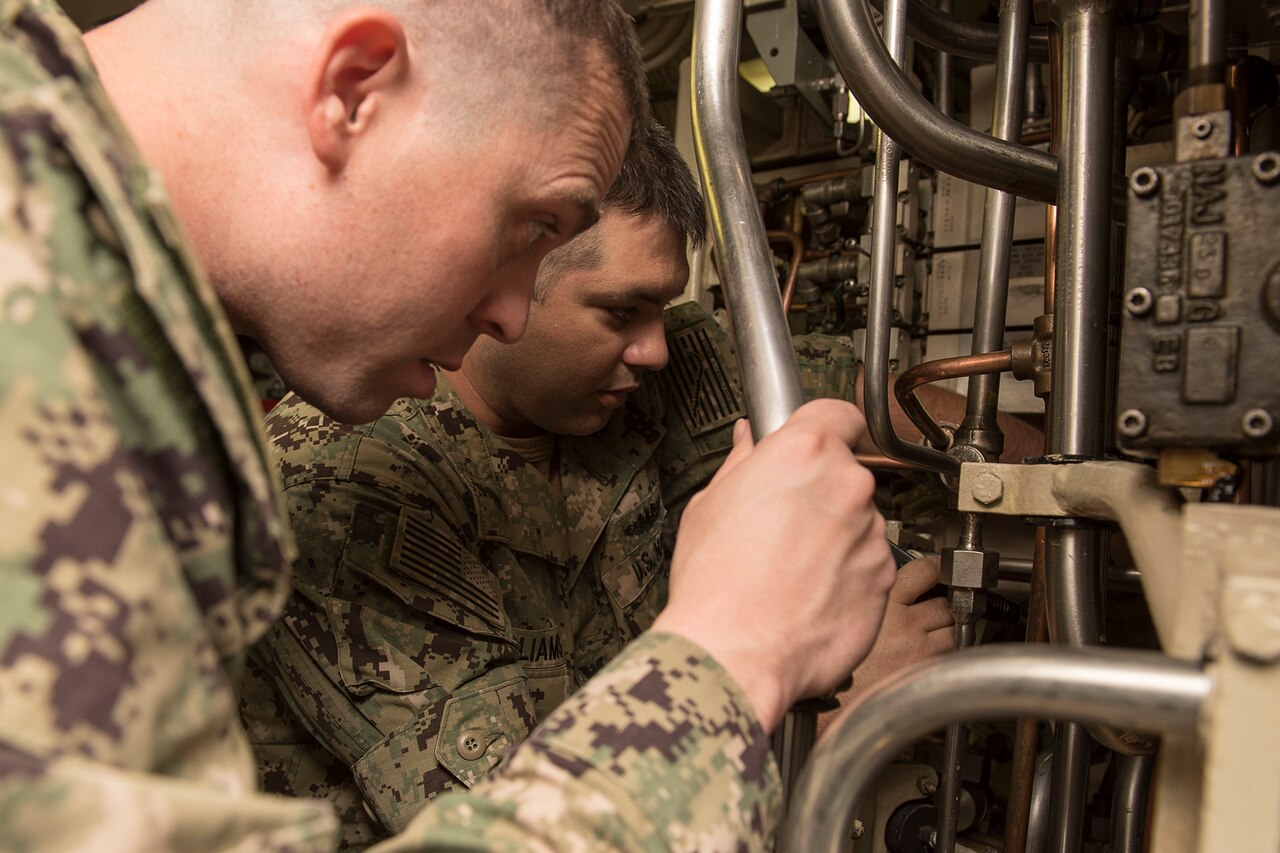 Two uniformed service members inspect equipment onboard a ship.