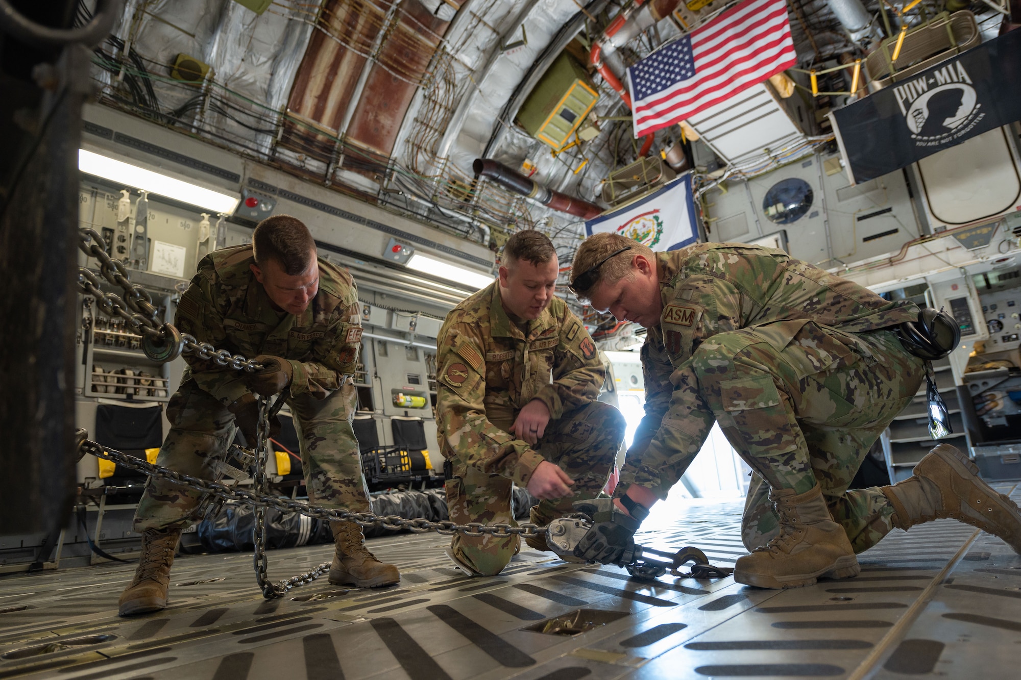 Three Airmen kneel in an aircraft and attach a chain to the cargo bay floor