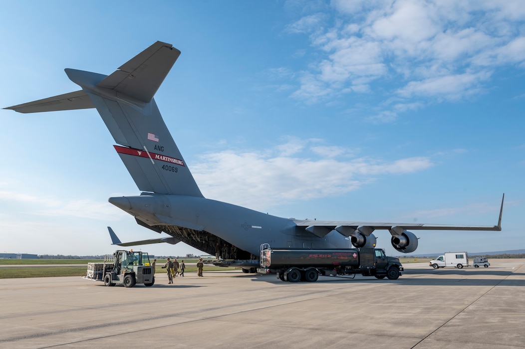 a cargo aircraft sits on a flightline with a fuel truck parked near it and a forklift carries a pallet towards the aircraft