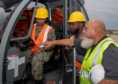 A female student in a working Navy uniform sits in the driver sear of a forklift. A male student and male instructor stand outside of the cab. the instructor is pointing at the control panel of the fork lift.