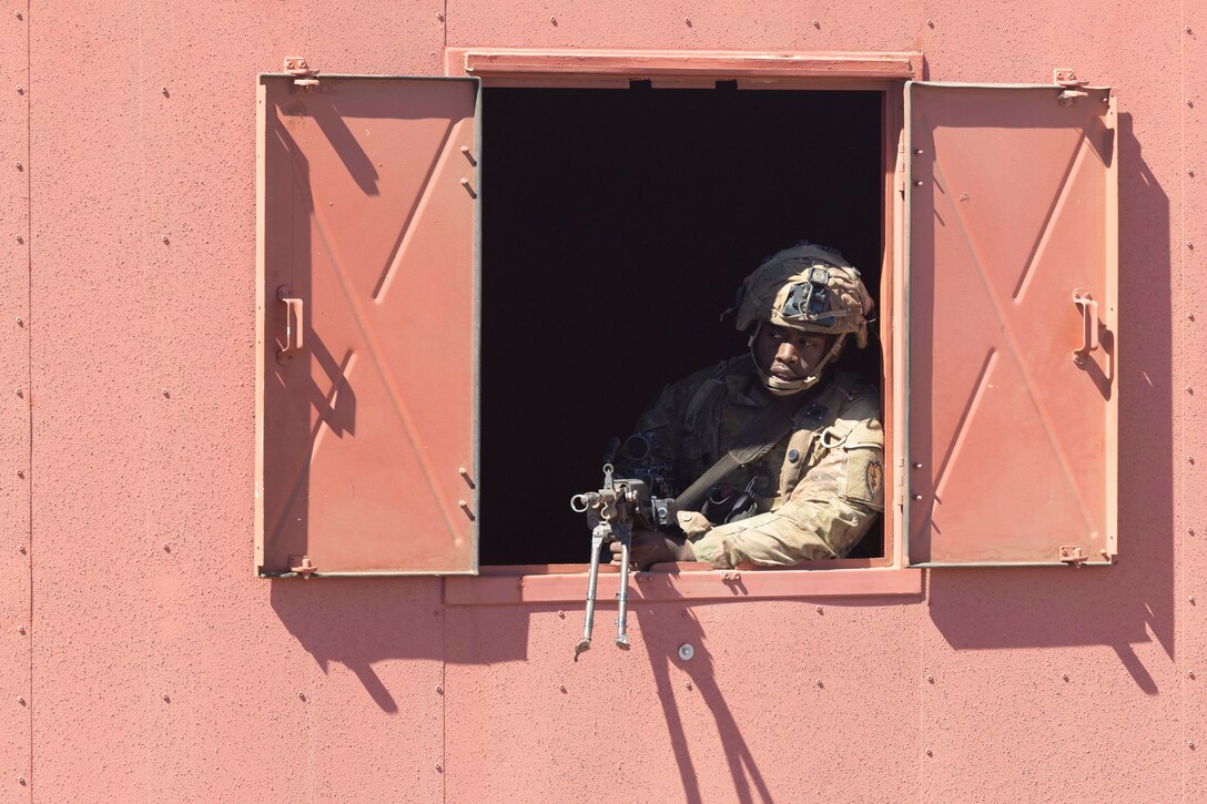 A soldier aims a weapon out of a window of a reddish building while standing watch.