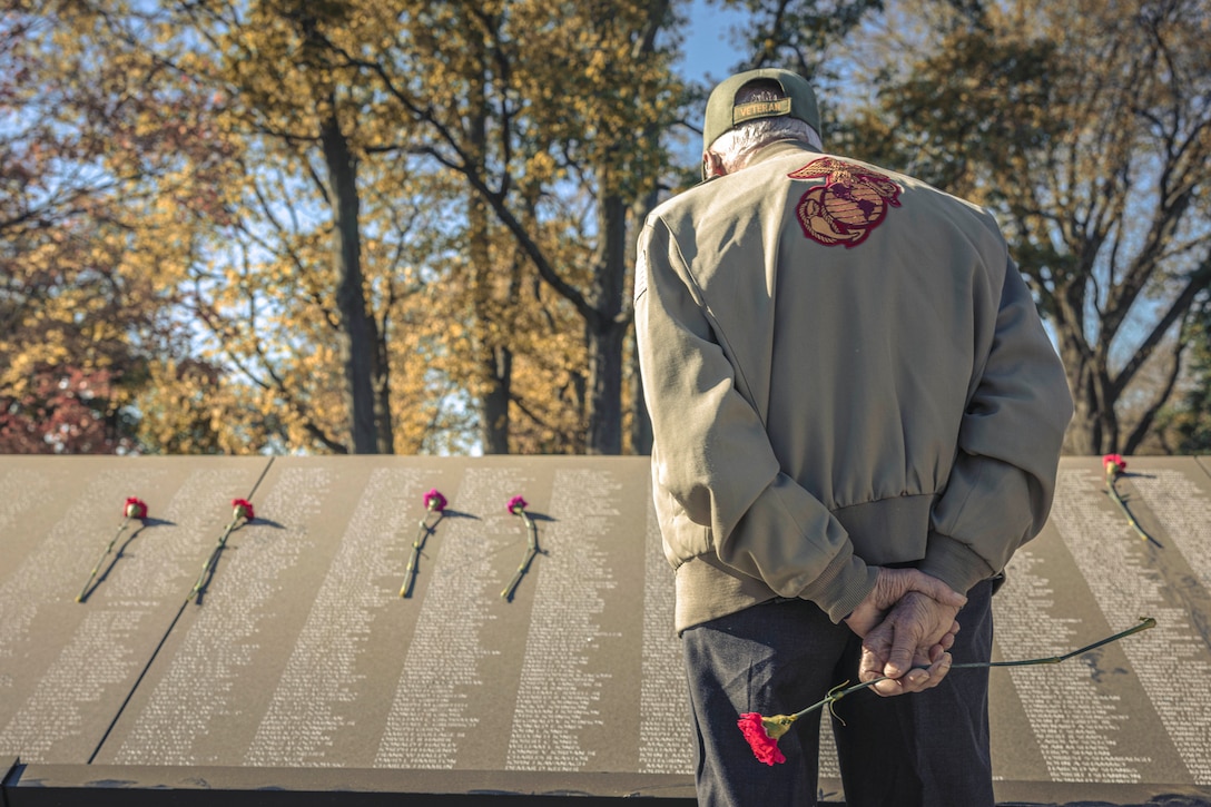 A veteran holds a rose behind their back while looking at a memorial of names with roses laid in between each row.