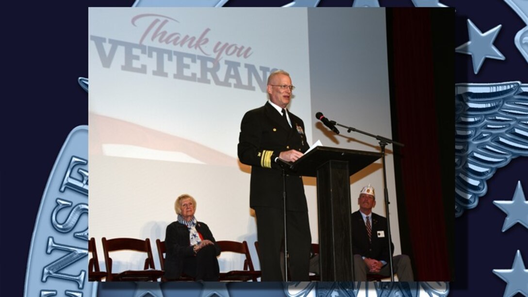 Photo is of a man in a Navy military uniform speaking from a podium during a Veterans Day ceremony.
