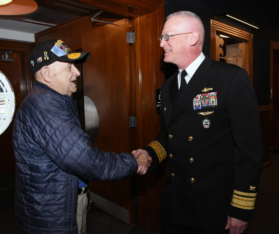 Photo is of two men standing shaking hands, one man in a Navy military uniform