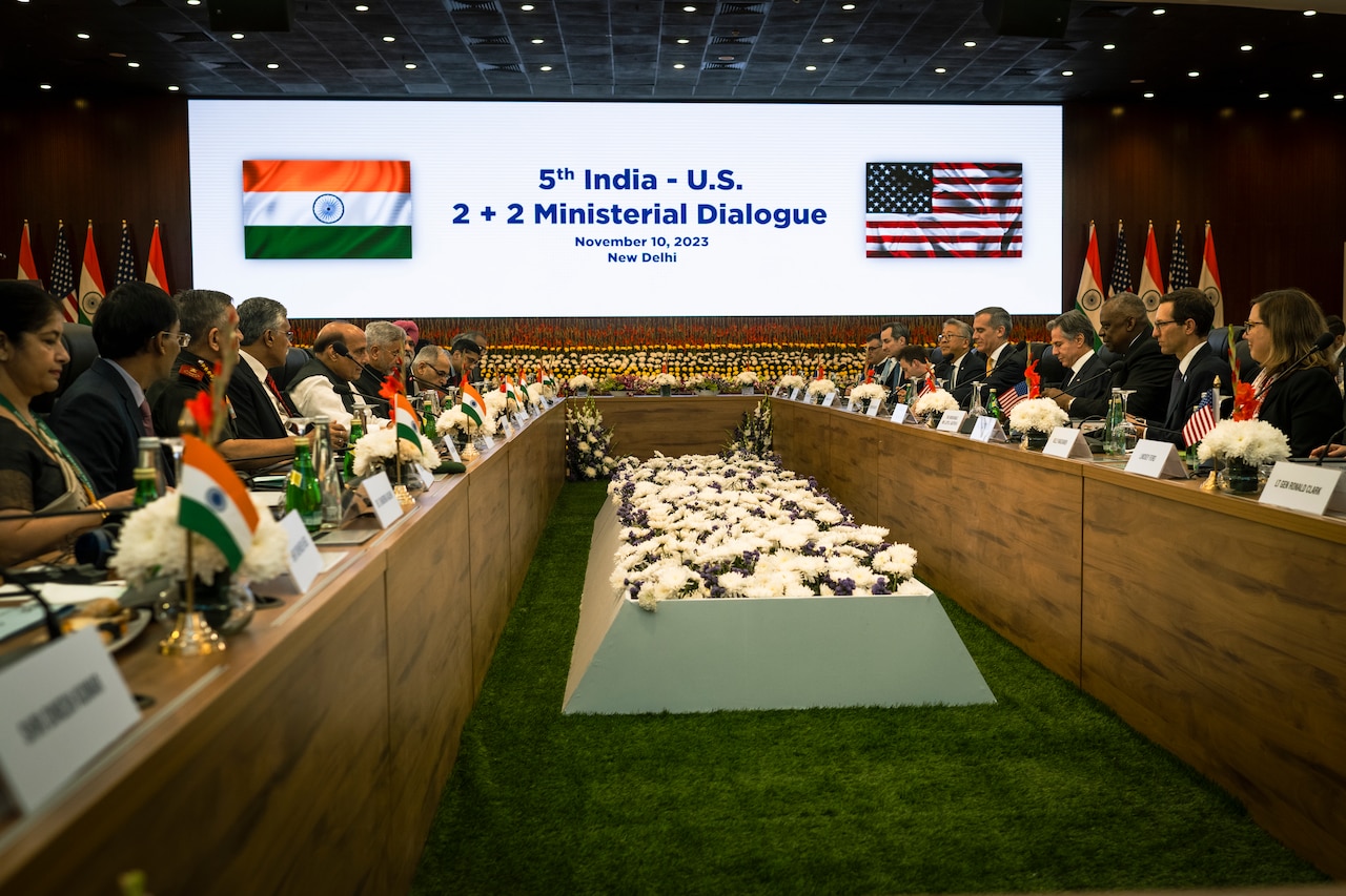U.S. and Indian officials gather at a table for meeting with a rectangular presentation in the background.