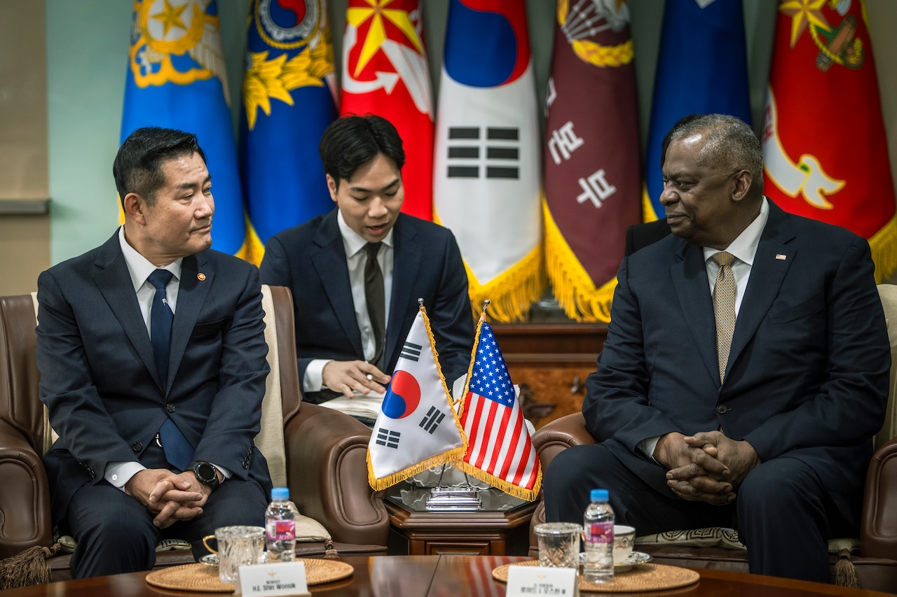 Two men in suits are seated next to a table displaying U.S. and South Korean flags.