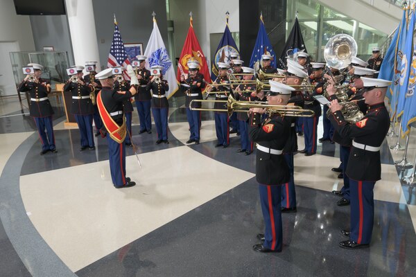 More than a dozen members of the Marine Corps Band Quantico play instruments in a semicircle in the McNamara Headquarters Complex cafeteria