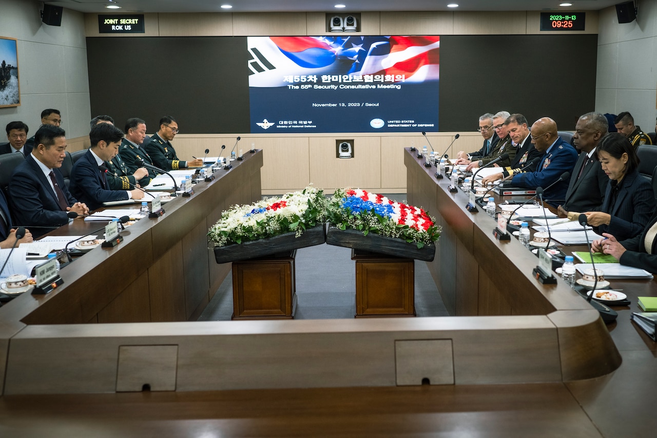Men and women wearing business attire and military uniforms are seated at a table with flower displays that depict the U.S. and South Korean flags.