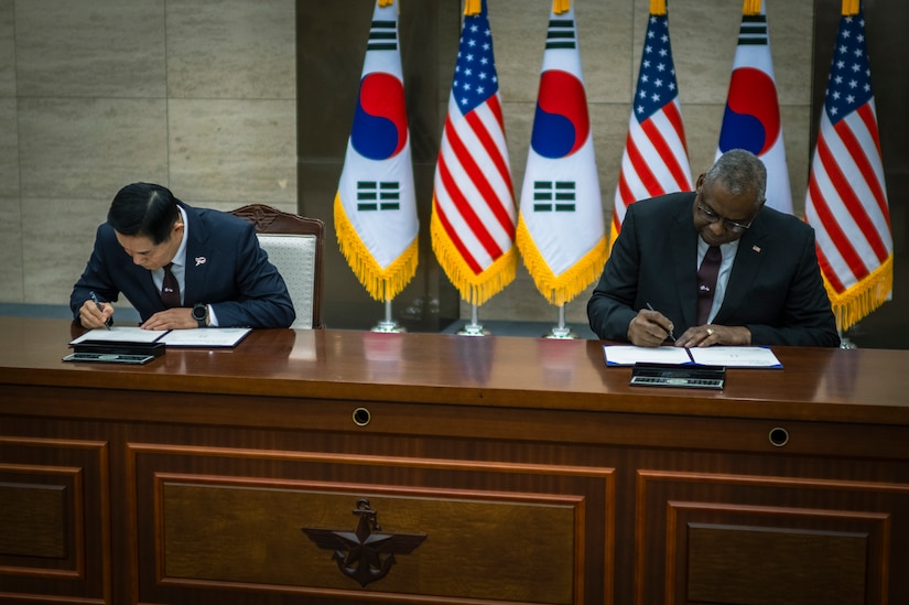Two men in suits sign documents while seated at a table with U.S. and South Korean flags in the background.