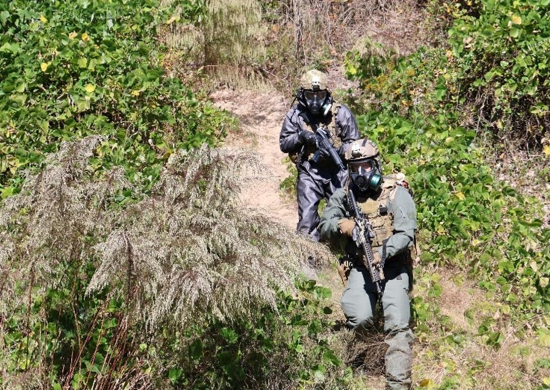 Two soldiers wearing CBRN protective gear walk down a hill during a training exercise with full coverage face masks, suites, gloves and boots.