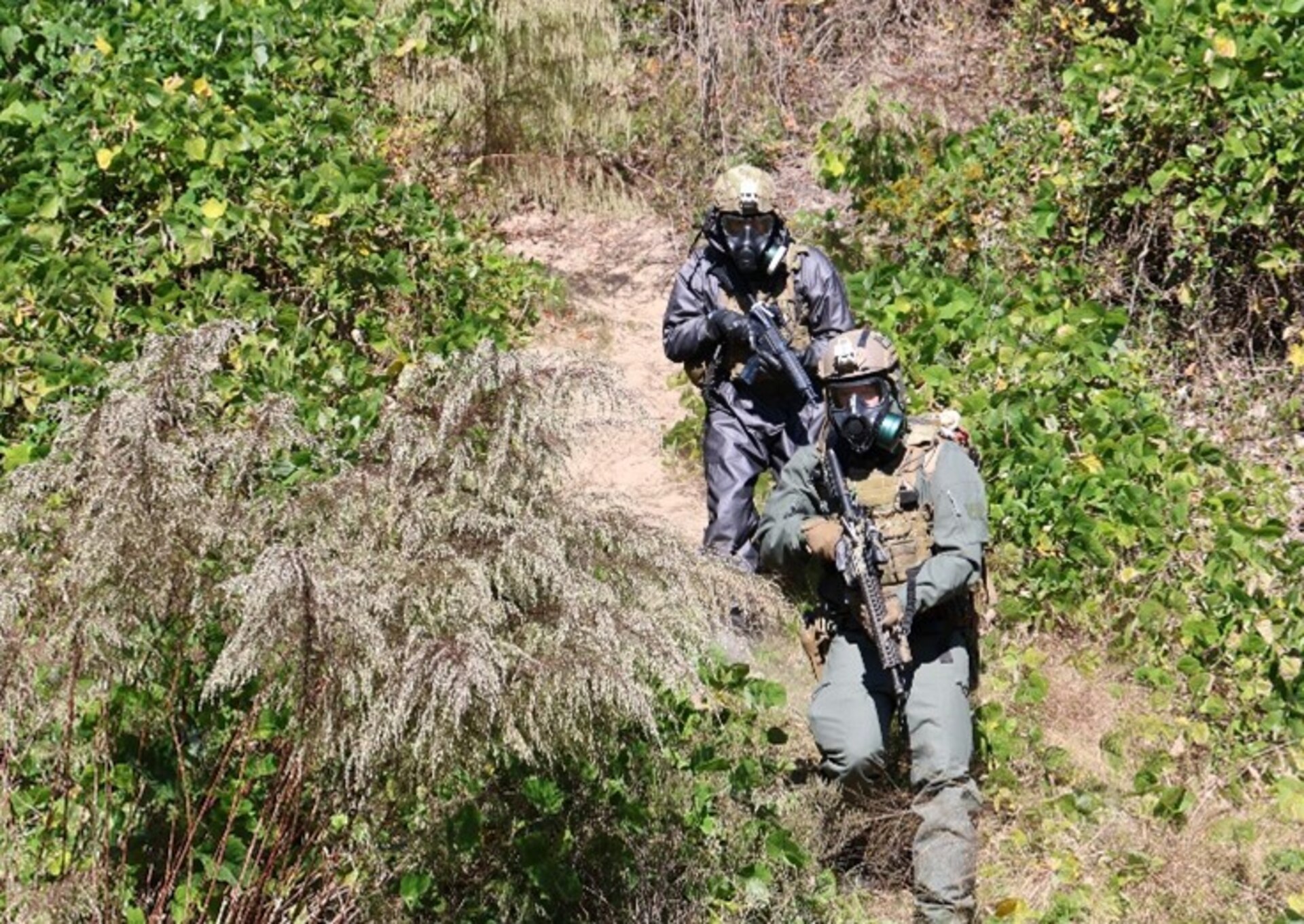 Two soldiers wearing CBRN protective gear walk down a hill during a training exercise with full coverage face masks, suites, gloves and boots.