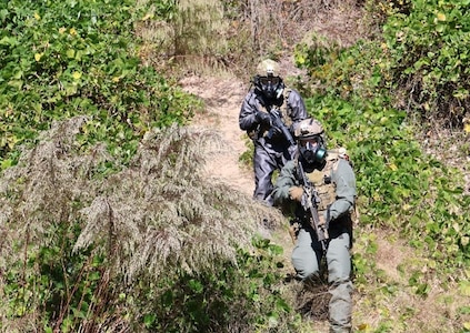 Two soldiers wearing CBRN protective gear walk down a hill during a training exercise with full coverage face masks, suites, gloves and boots.