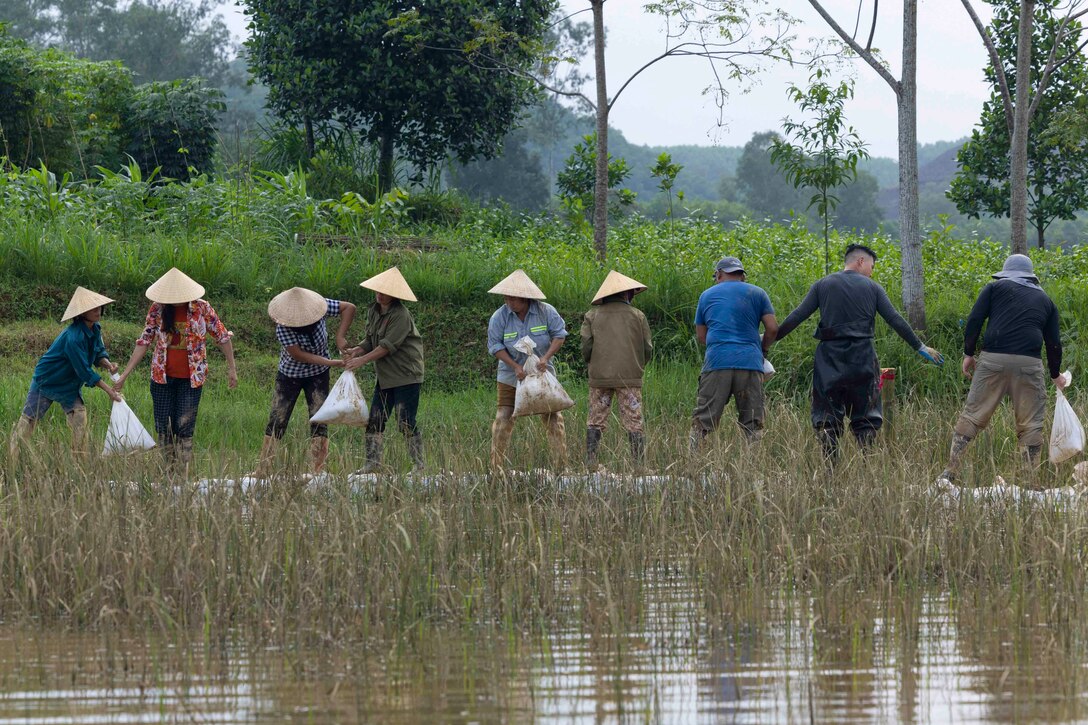 People stand in a line in a wet grassland while handing bags to each other.