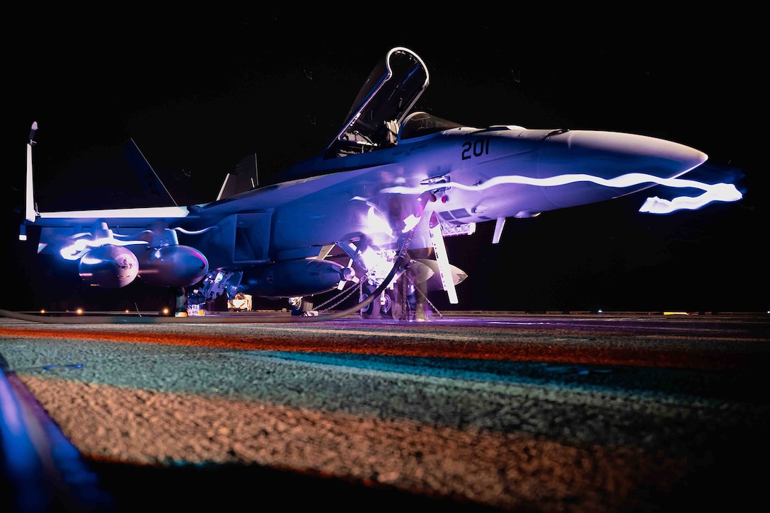 A sailor connects a fuel hose to a military aircraft at night. The photo features slow shutter lights across the front of the aircraft.