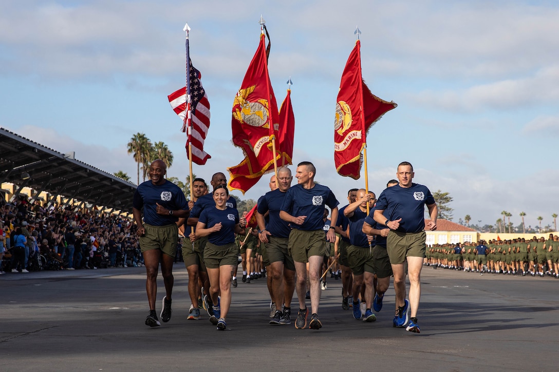 A group of Marines run as a crowd looks on. Four of the Marines are holding flags as they run.