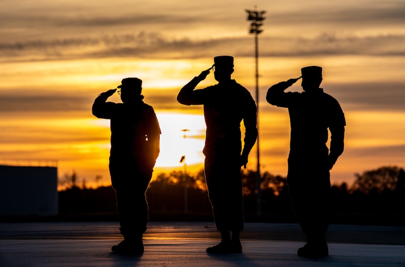 Three people salute on an airfield as the sun sets behind them.
