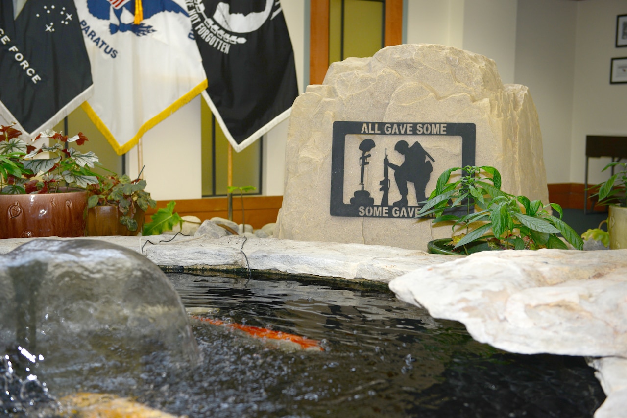 A water fountain bubbles near flags and a rocky monument that says, “All Gave Some, Some Gave All.”