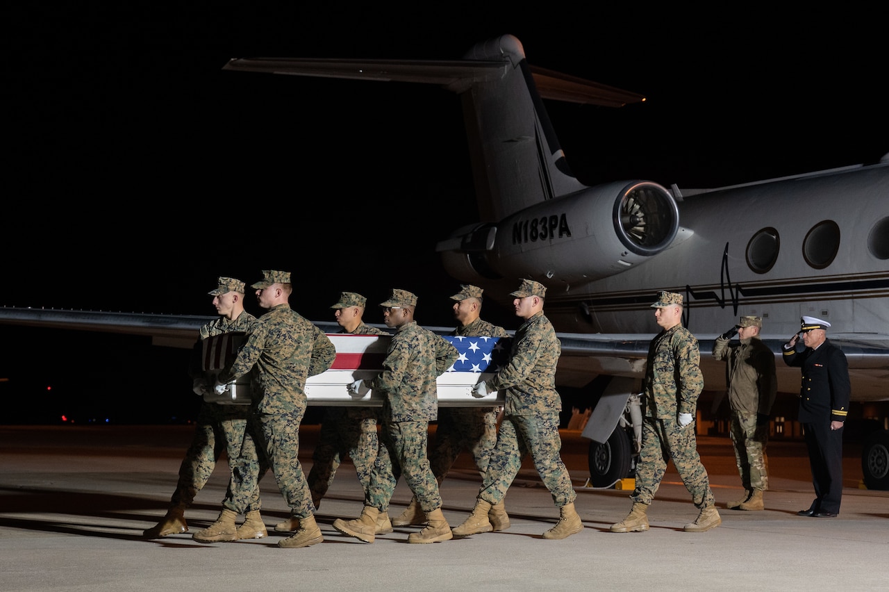 A team of men carries a transfer case holding the remains of a man. Two men in the background near an aircraft salute.