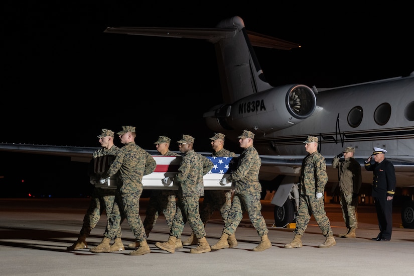 A team of men carries a transfer case holding the remains of a man. Two men in the background near an aircraft salute.
