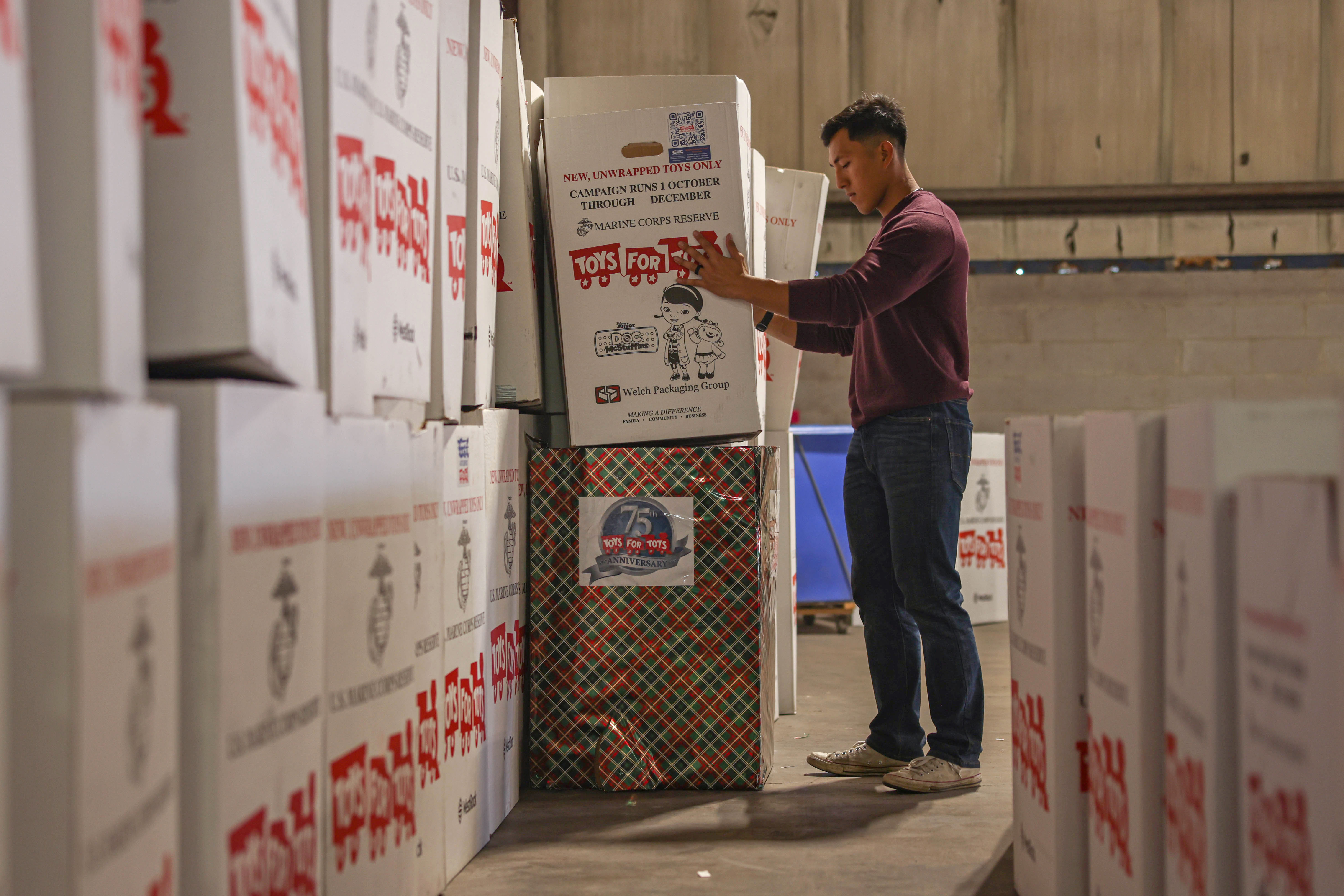 A Marine handles a large white box with “TOYS FOR TOTS” written on it in a warehouse-type space filled with similar boxes.