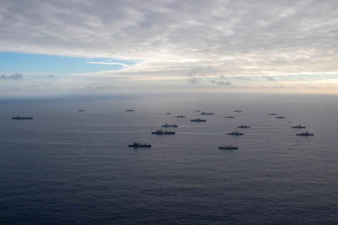 A wide shot of U.S., Japanese, Australian and Canadian naval ships sailing in formation under a cloudy sky as the sun peeks out on the horizon.