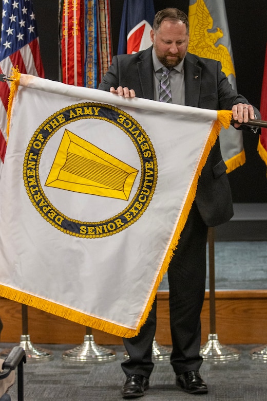 Ryan A. Busby, U.S. Army Financial Management Command deputy to the commanding general, unfurls his U.S. Army Senior Executive Service flag during his SES induction ceremony at the Maj. Gen. Emmett J. Bean Federal Center in Indianapolis Nov. 2, 2023. The SES insignia is a keystone, which is a center stone that holds all the stones of an arch in place, and this represents the critical role of the SES as a central coordinating point between government's political leadership and the line workers. (U.S Army photo by Mark R. W. Orders-Woempner)