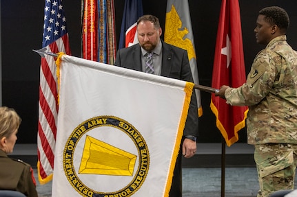 Maj. Dane Wright, U.S. Army Financial Management Command Operations chief of plans, helps Ryan A. Busby, USAFMCOM deputy to the commanding general, unfurl his U.S. Army Senior Executive Service flag during Busby’s SES induction ceremony at the Maj. Gen. Emmett J. Bean Federal Center in Indianapolis Nov. 2, 2023. The SES insignia is a keystone, which is a center stone that holds all the stones of an arch in place, and this represents the critical role of the SES as a central coordinating point between government's political leadership and the line workers. (U.S Army photo by Mark R. W. Orders-Woempner)