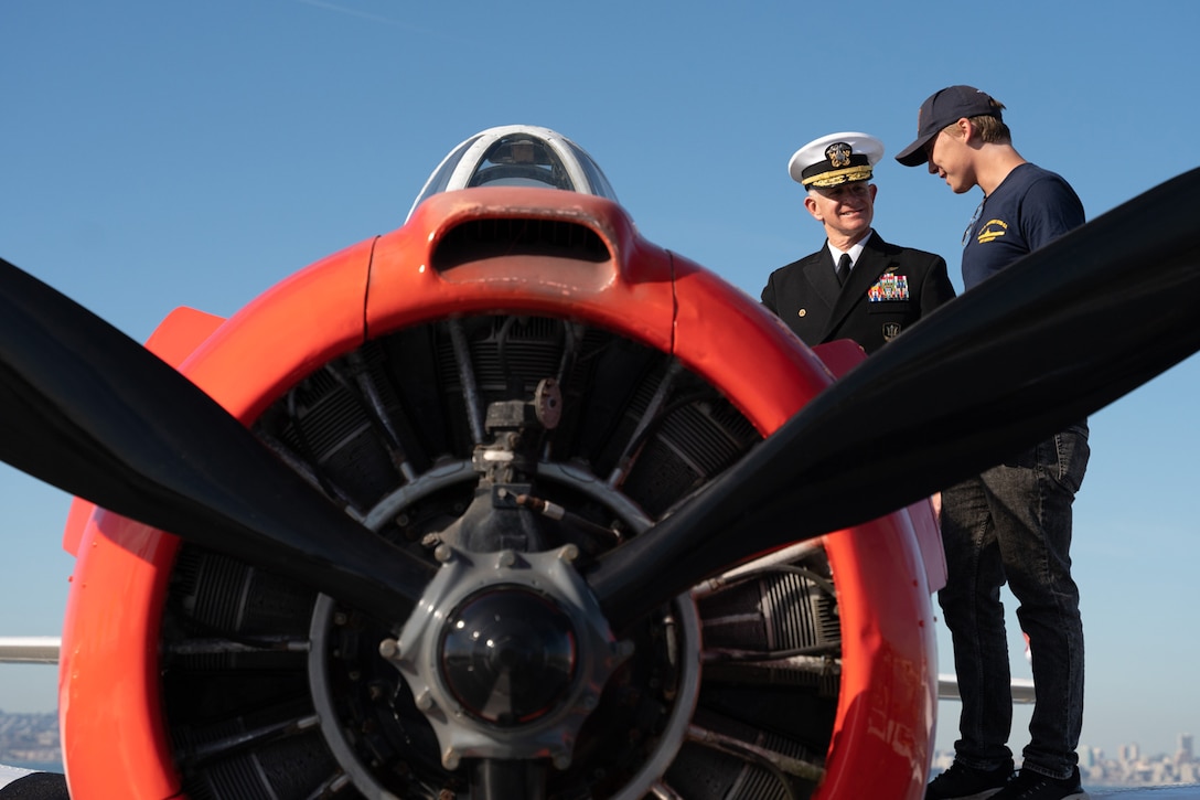 Two service members stand next to an aircraft. The aircraft’s propellers are shown close-up.