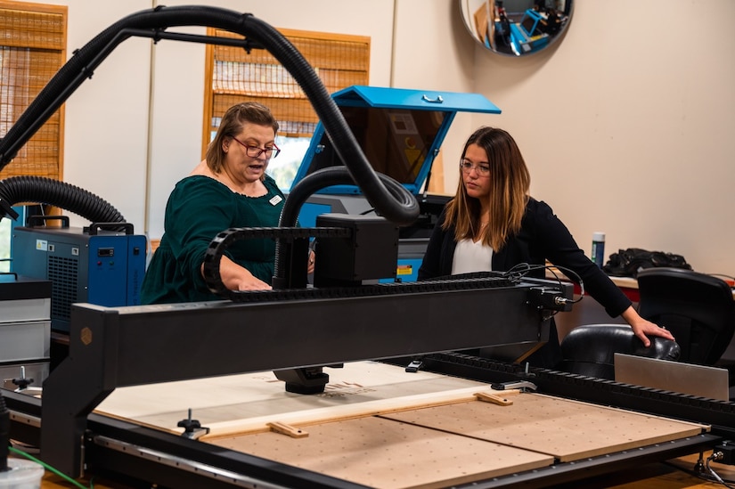 U.S. Air Force Lana Simmons, Bateman library technician and innovation lab manager, and Ashley Jellison, library director, use a computer numerical control router to engrave a sign for the innovation lab at Joint Base Langley-Eustis, Virginia, Nov 6, 2023. The lab was created for anyone with access to the Bateman library. (U.S. Air Force photo by Airman 1st Class Ian Sullens)