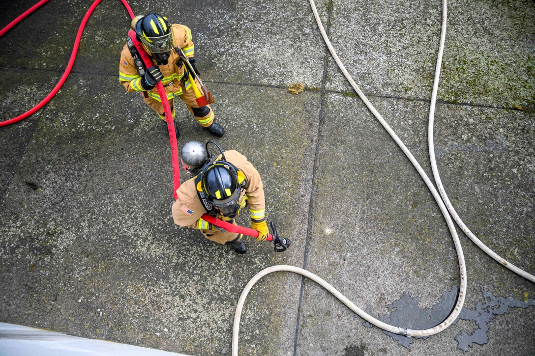Two firefighters carrying a red hose walk near a white hose laying on the ground as seen from above.
