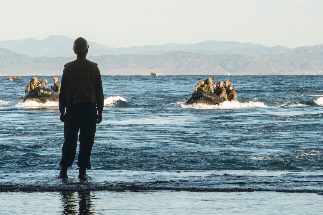 A service member stands watch on shore while U.S. and British marines transit a body of water in small boats with mountains in the background.