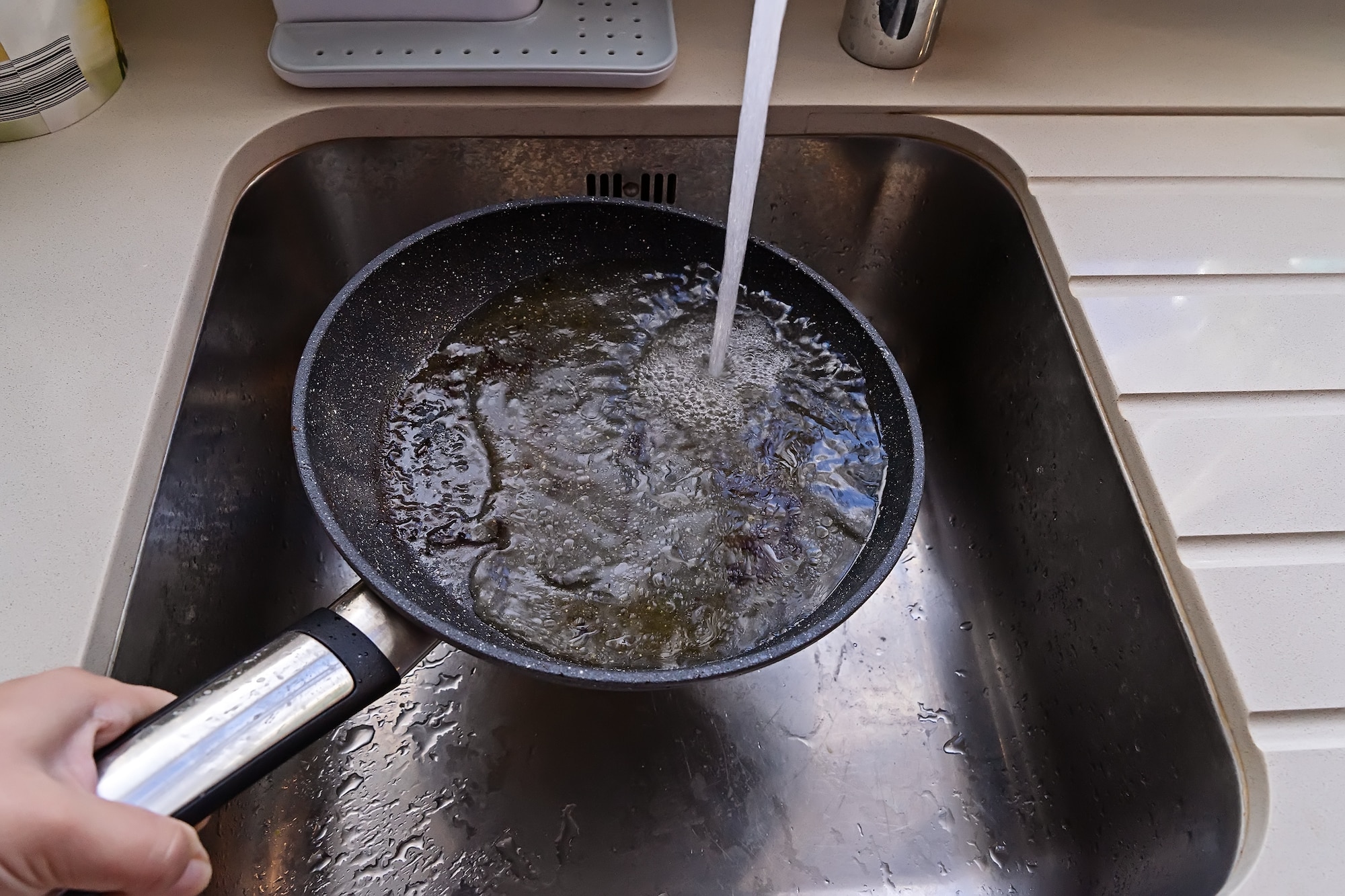 Hand holding a pan over a sink while faucet is running water over it.