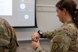 a woman service member stands and looks at her wrist, equipped with a wrist-worn device that looks like a smart watch.