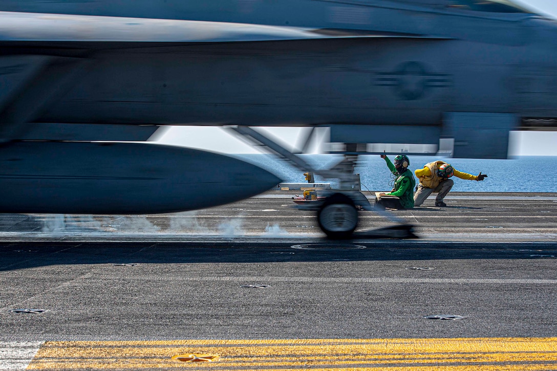 Two sailors on a flight deck of a ship signal for the launch of an F/A-18F Super Hornet fighter jet.