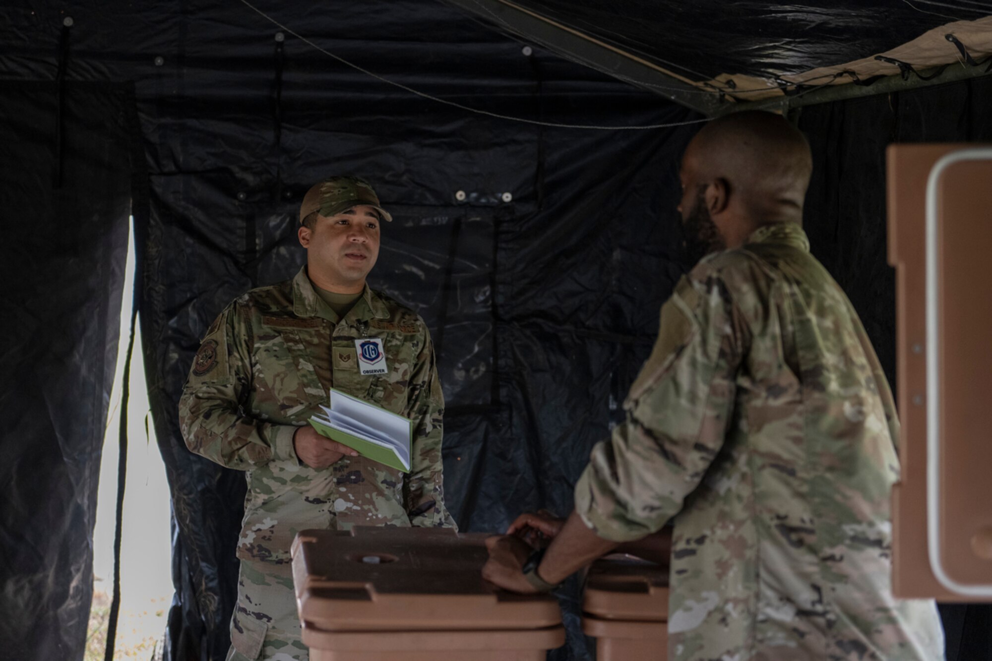 U.S. Air Force Staff Sgt. Victor Santacruz-Mitchell, 23rd Wing Personnel Support for Contingency Operations member, left, asks Senior Airman Derrick Brock, 23rd Force Support Squadron shift lead, questions for an evaluation during exercise Mosaic Tiger 24-1 at Avon Park Air Force Range, Florida, Nov. 14, 2023. As a Services observer, Santacruz-Mitchell takes notes and evaluates how well 23rd FSS Airmen conduct their operations in a contingency location. (U.S. Air Force photo by Senior Airman Rachel Coates)